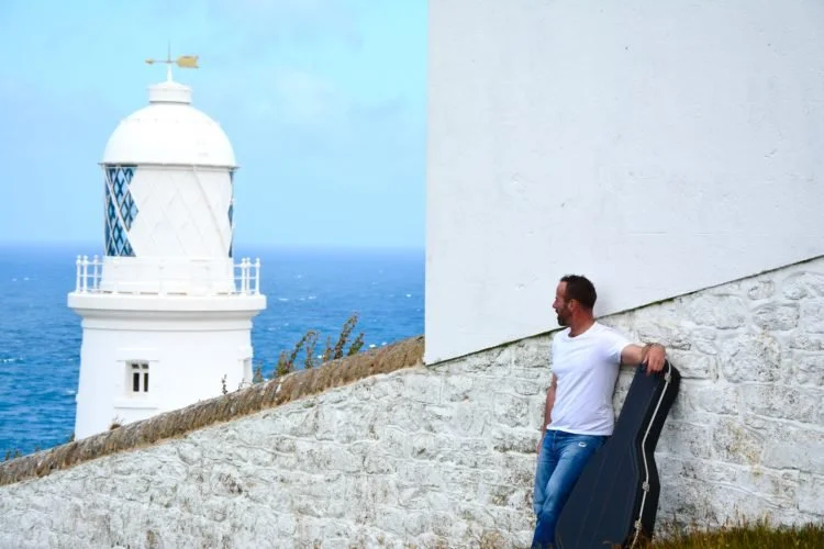 Man leaning against a white stone wall with a small white lighthouse in the background, holding a guitar case.