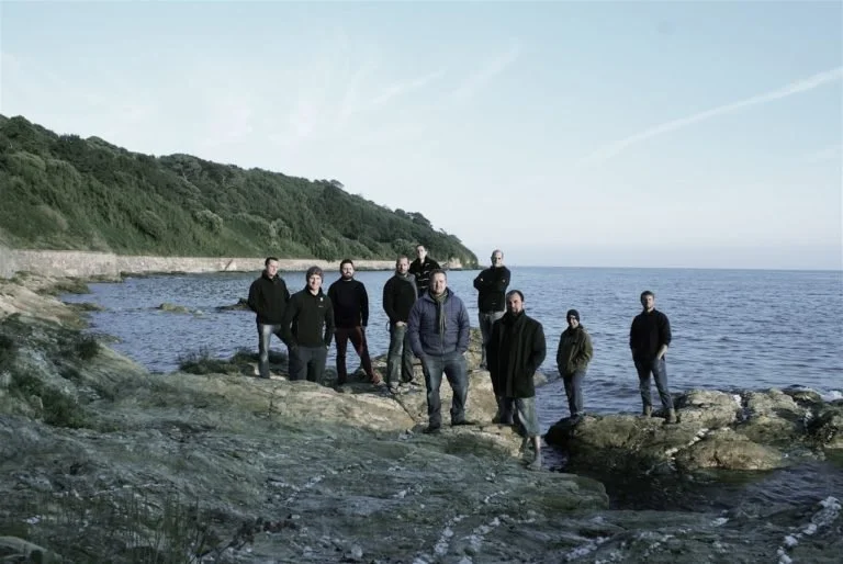 Ten people standing on rocks by the ocean with a wooded hillside in the background.