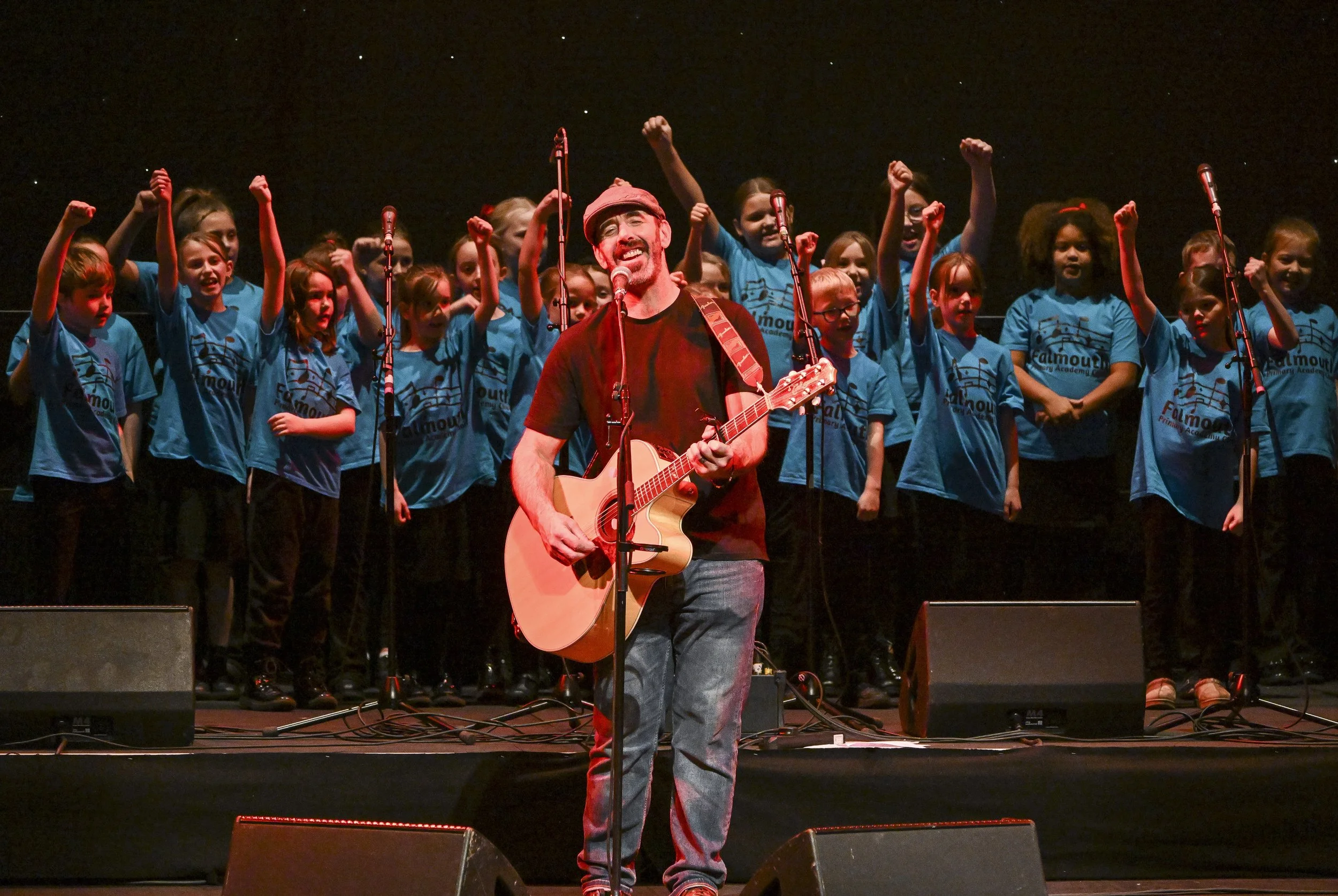 A man singing and playing guitar on stage, surrounded by a children's choir in blue t-shirts, with their arms raised in the air.