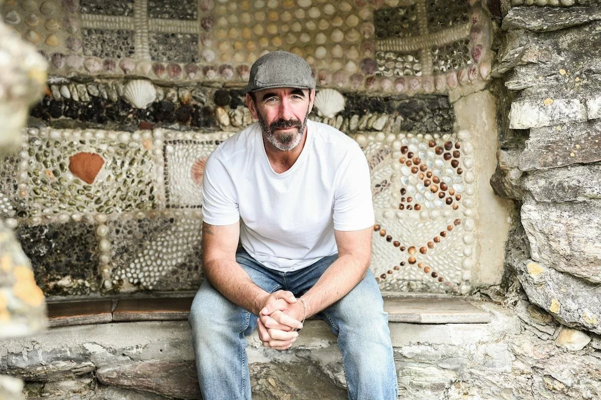 A man with a beard, wearing a gray cap and white t-shirt, sitting on a ledge in front of a wall decorated with seashell mosaics.
