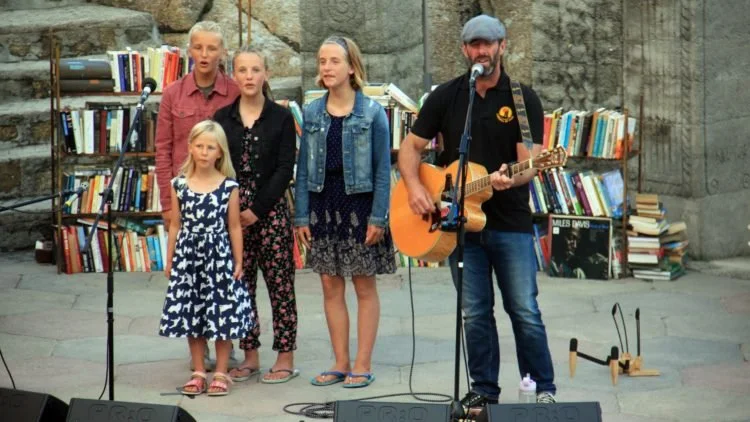 A man with a beard playing guitar and singing into a microphone while four women and a girl sing behind him, set against stone wall and bookshelf background at an outdoor event.