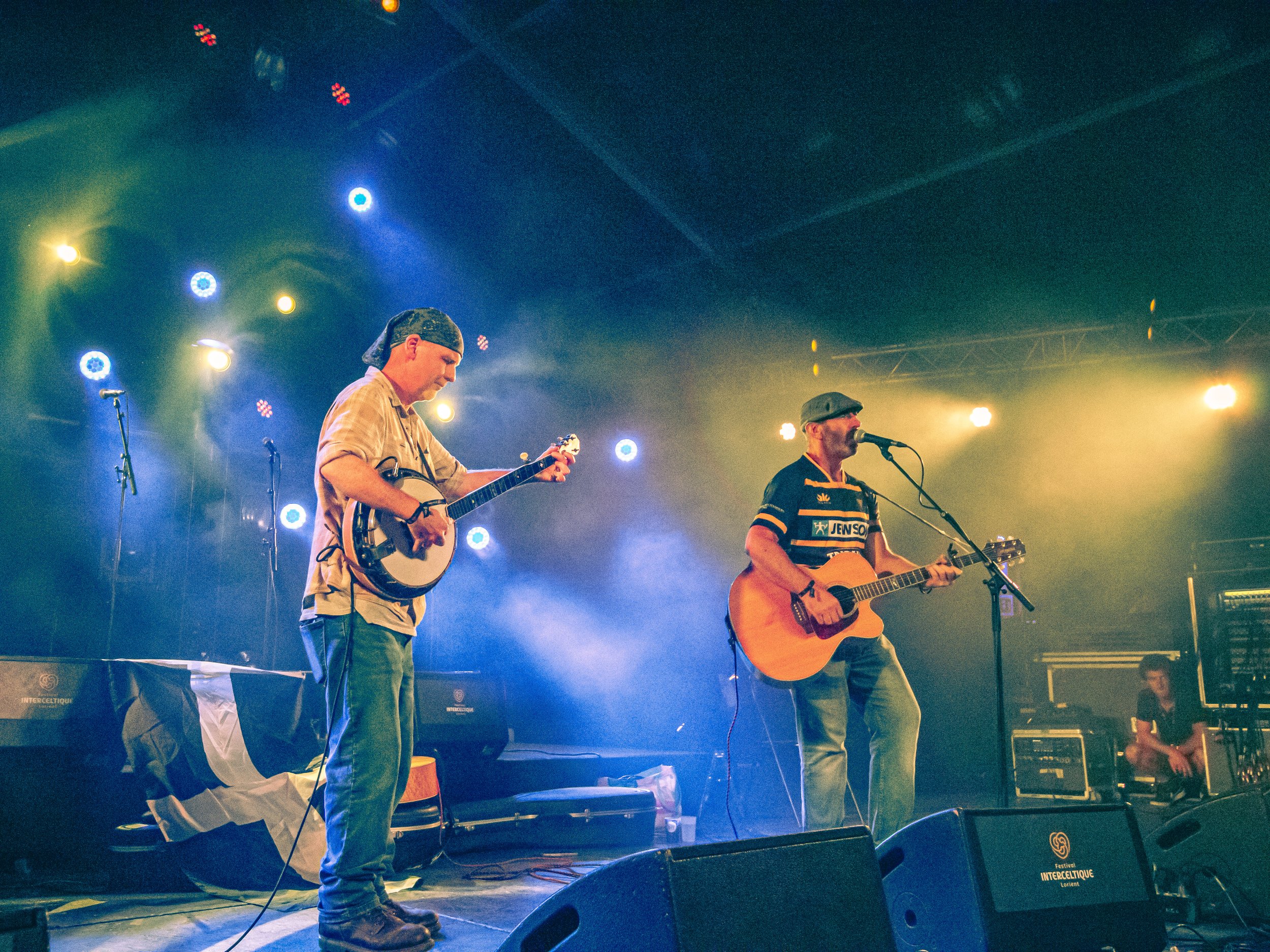 Two musicians playing guitars on stage with colorful lighting, one wearing a beige shirt and bandana, the other in a striped shirt and flat cap, with a person in the background operating sound equipment.