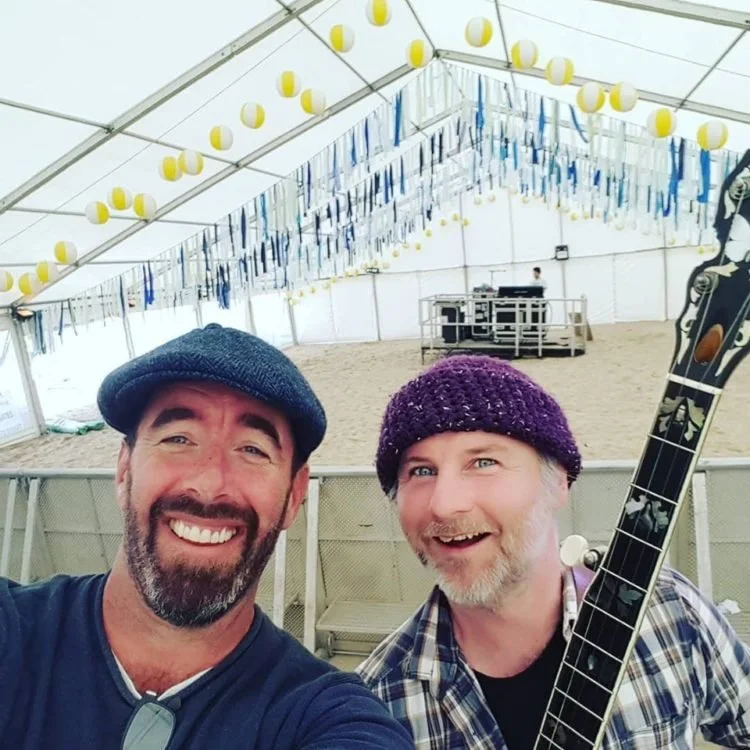 Two smiling men wearing hats inside a decorated tent, one holding a guitar, with a stage and DJ equipment in the background.