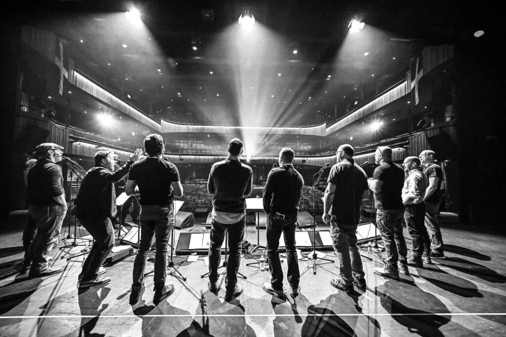 A group of people on a stage in a theater, with spotlights shining down on them, preparing for a performance or rehearsal.