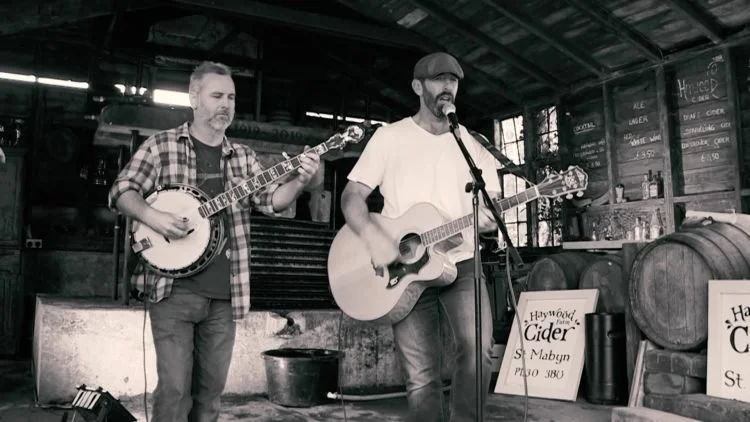 Two musicians playing guitars and singing in a rustic indoor setting, with barrels and a chalkboard menu in the background.