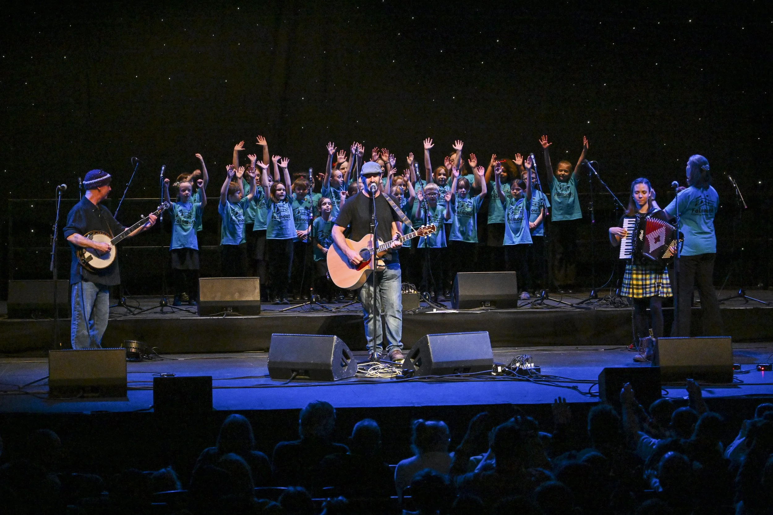 Group of children on stage, some with their hands raised, performing with musical instruments with audience watching in a darkened theater.