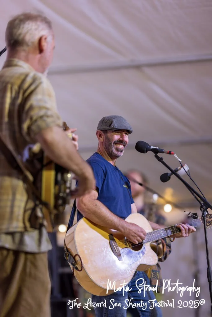 Two men performing music at Lizard Sea Shanty Festival 2025, one playing a guitar and singing, the other with a bass guitar, both in casual clothing inside a tent.