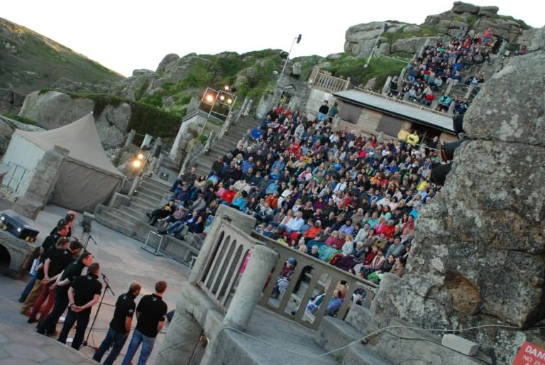 An amphitheater built into rocky hillside with a large audience seated on tiered stone steps and a small group of people standing in front.