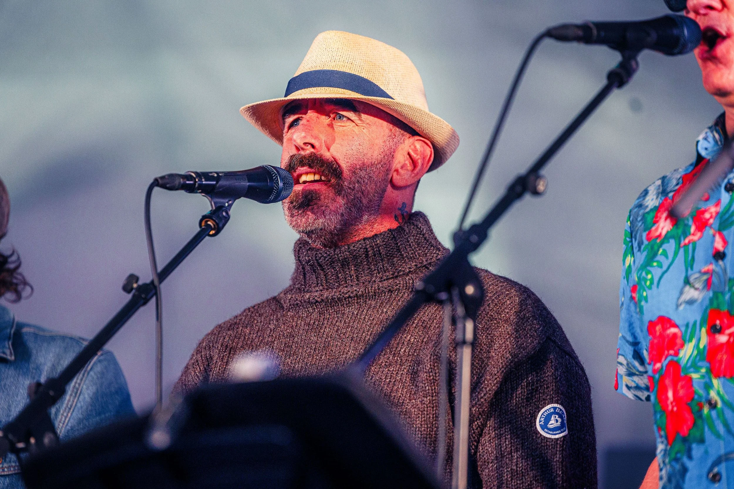 A man with a beard and mustache, wearing a tan fedora hat and a dark brown knit turtleneck sweater, is singing or speaking into a microphone during a performance. Partially visible on his left and right are other performers with microphones. The back