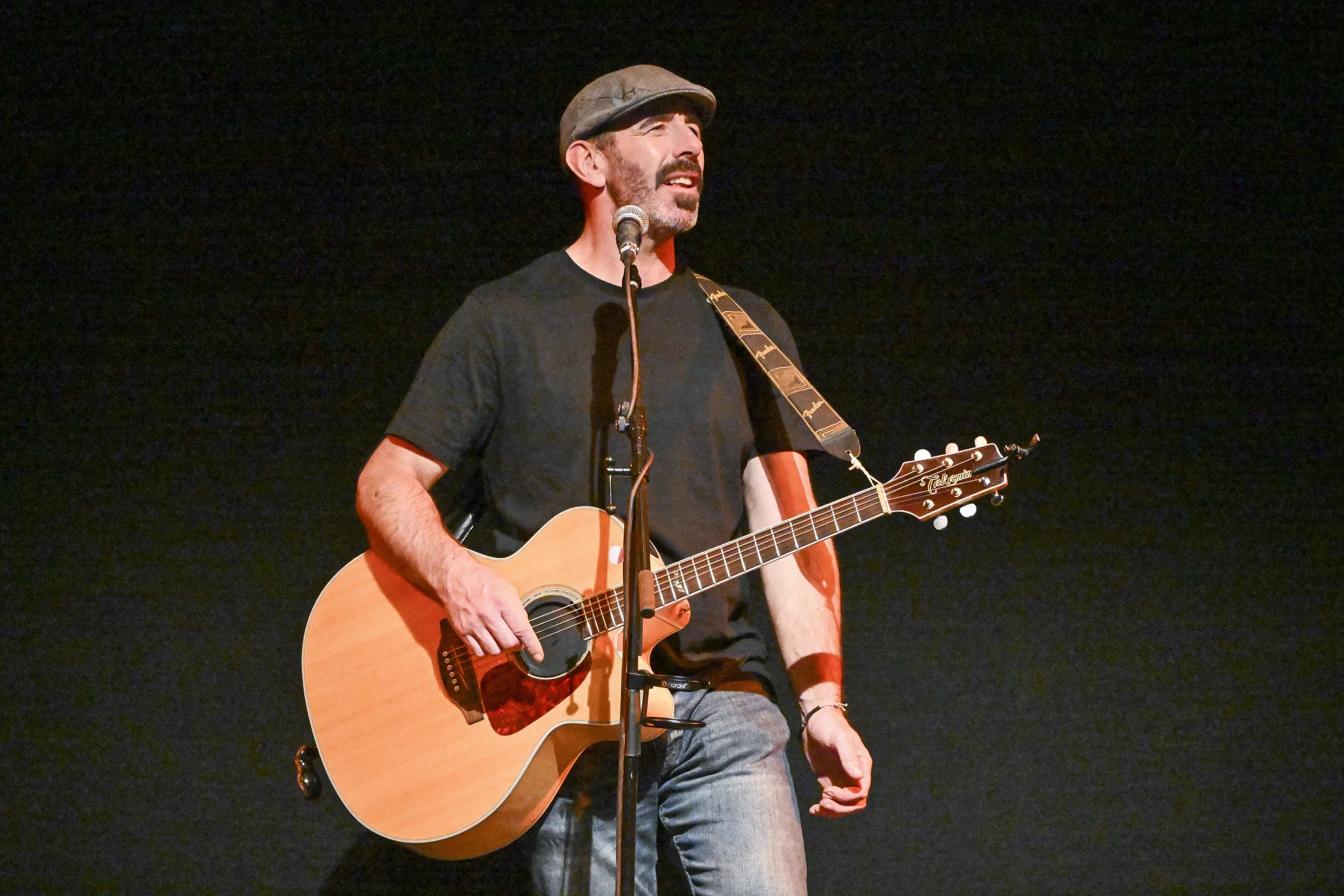 Man singing into a microphone while playing an acoustic guitar on a dark stage.