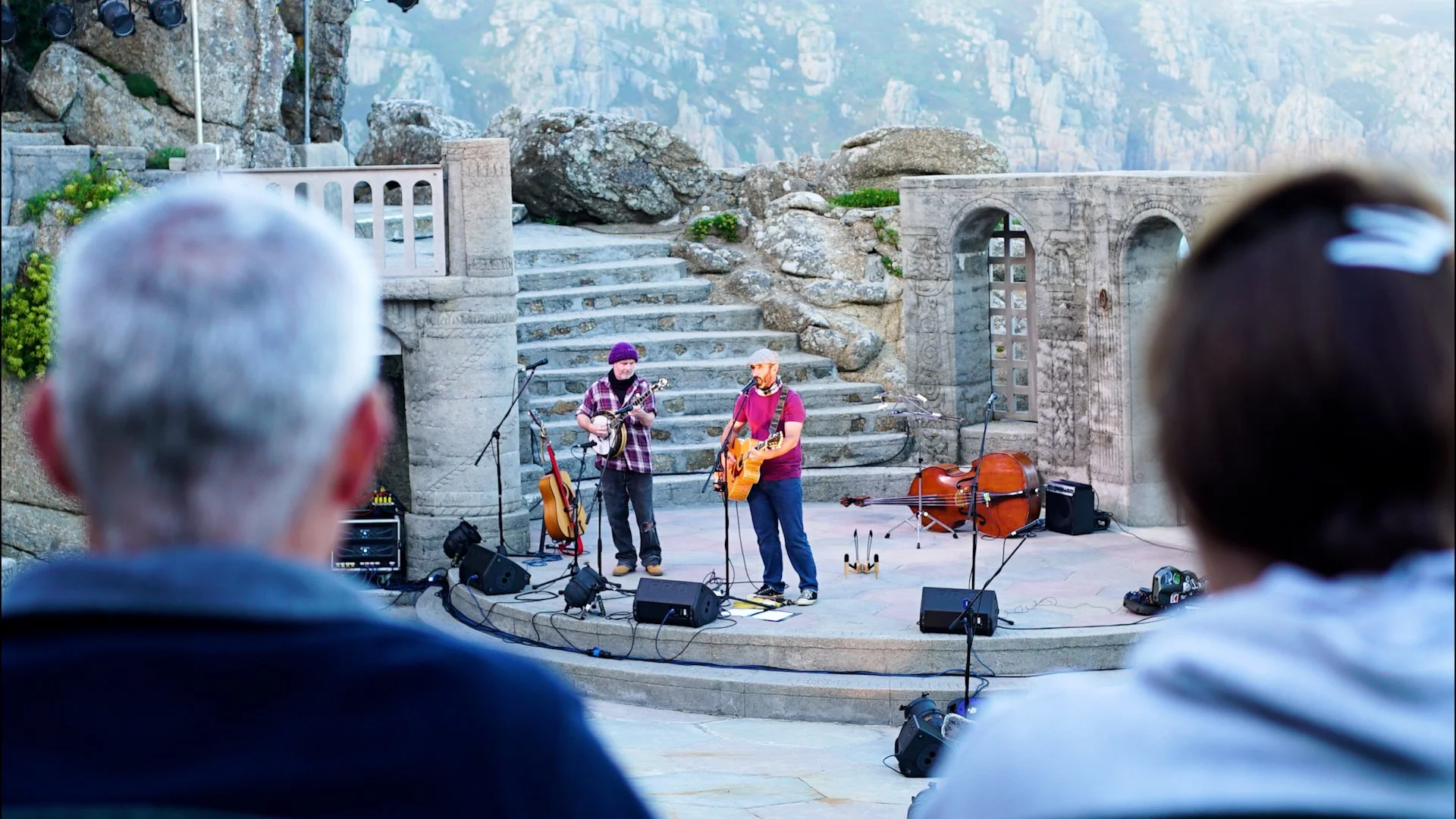 Two musicians perform on an outdoor stage with stone steps and ancient stone walls, overlooking a mountainous landscape, as two people in the foreground watch.