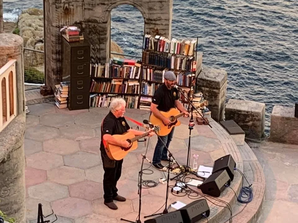 Two musicians playing acoustic guitars on a stone stage near the water, with bookshelves and a stone arch in the background.