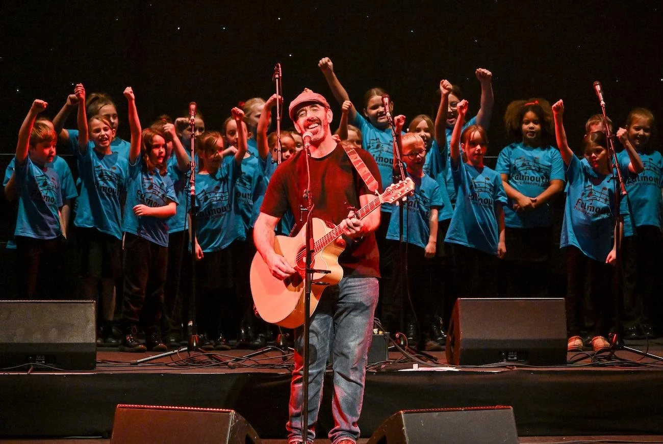 A man with a guitar performing on stage with a children's choir in blue T-shirts behind him, some children raising their fists.