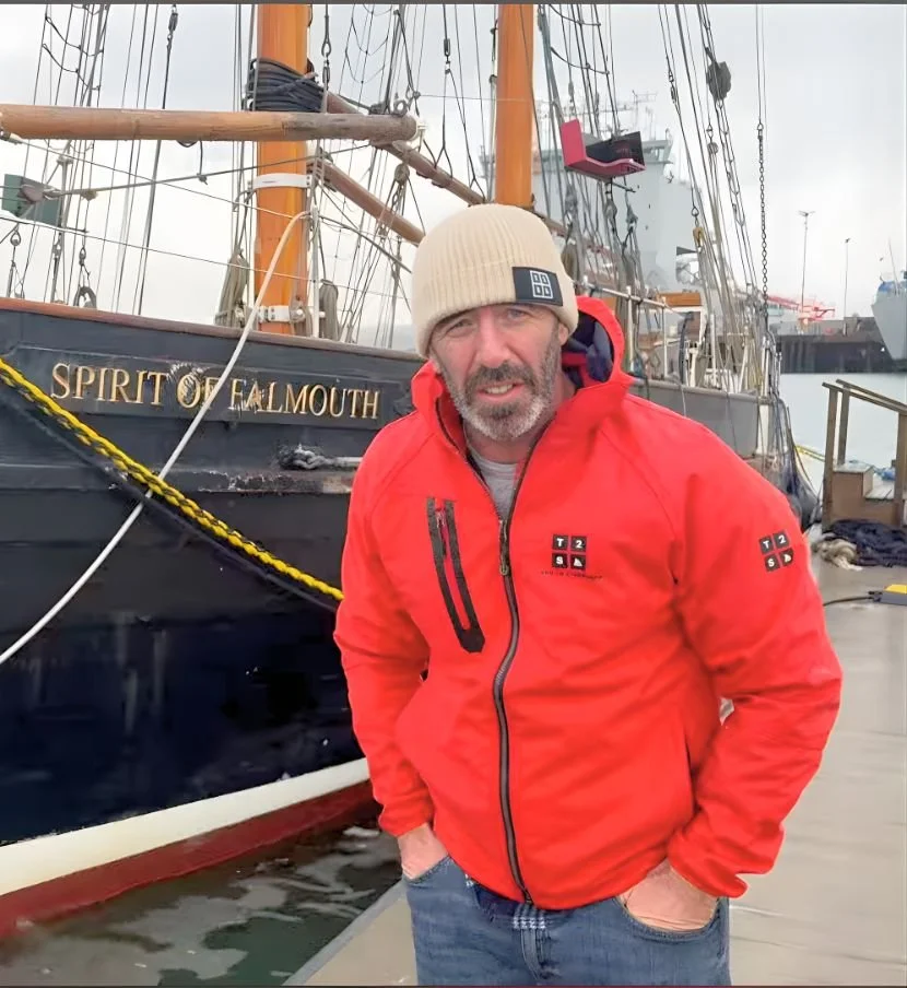 A man with a beard wearing a beige knit cap and red jacket standing on a dock next to a tall sailing ship named Spirit of Falmouth.