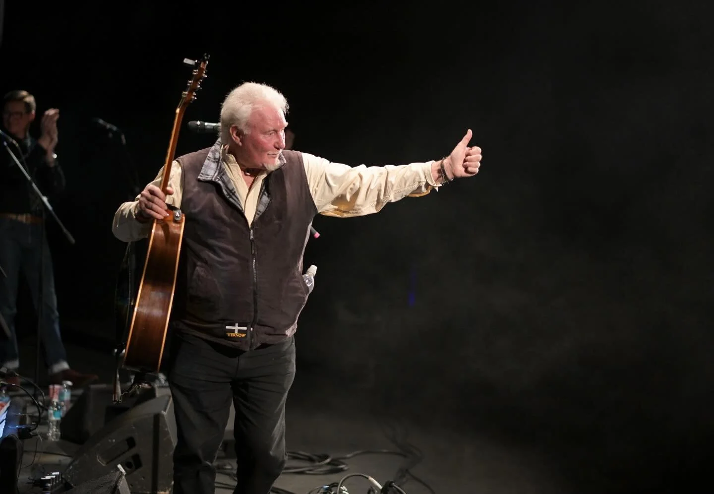 An elderly man with white hair giving a thumbs-up while holding a guitar on stage during a performance.
