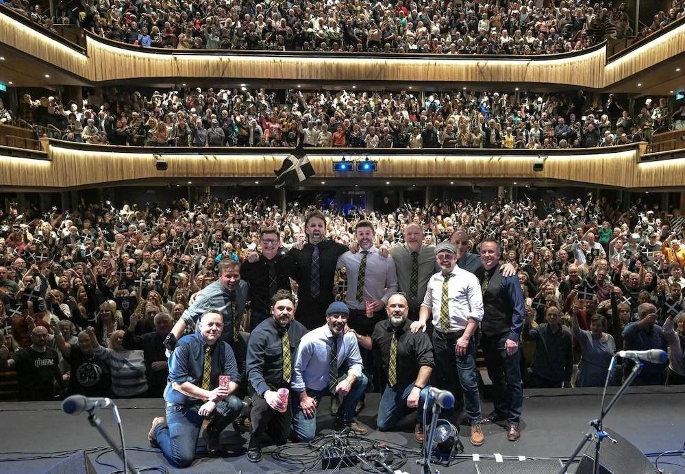 A large concert hall filled with a crowd of people, some holding small flags. In the foreground, a group of ten men poses on stage, some kneeling and some standing, all smiling. The audience appears excited and engaged.