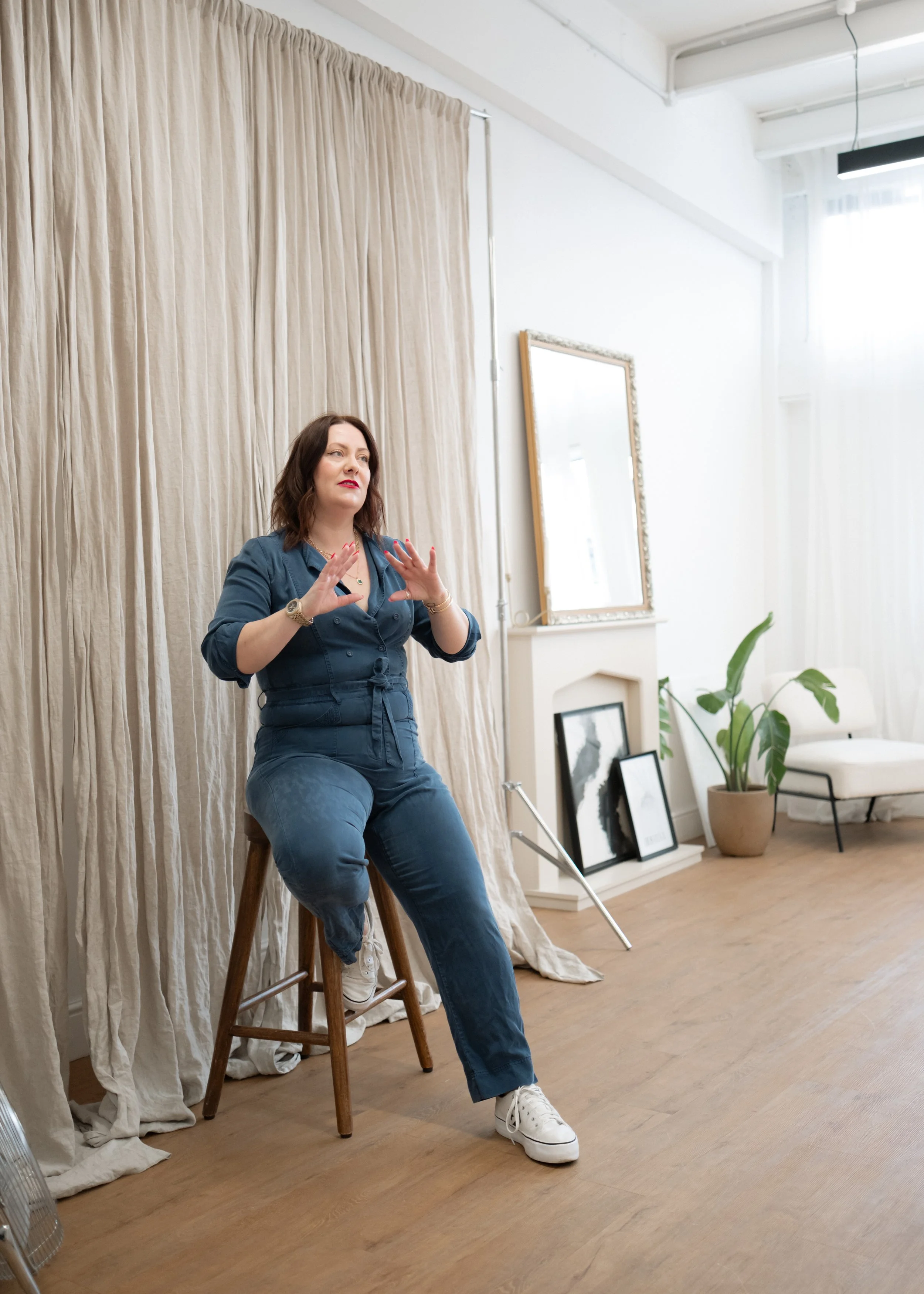 A woman with brown hair and red lipstick sitting on a stool in a room with white walls, beige curtains, and wooden flooring. She is wearing a blue outfit, white sneakers, and appears to be gesturing with her hands.