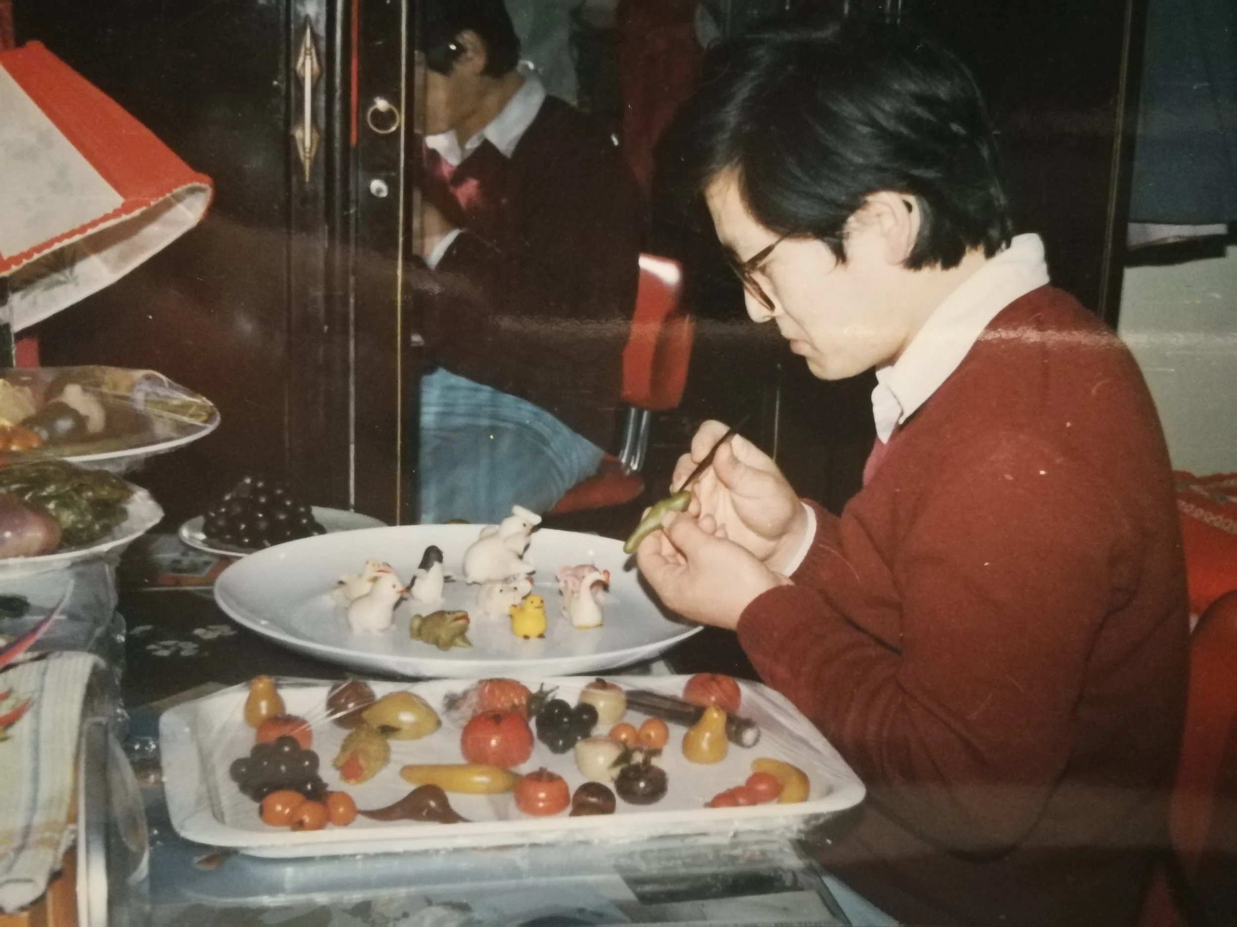 A person sitting at a table decorating small figurines with a paintbrush, surrounded by various confections and chocolates on plates and trays.