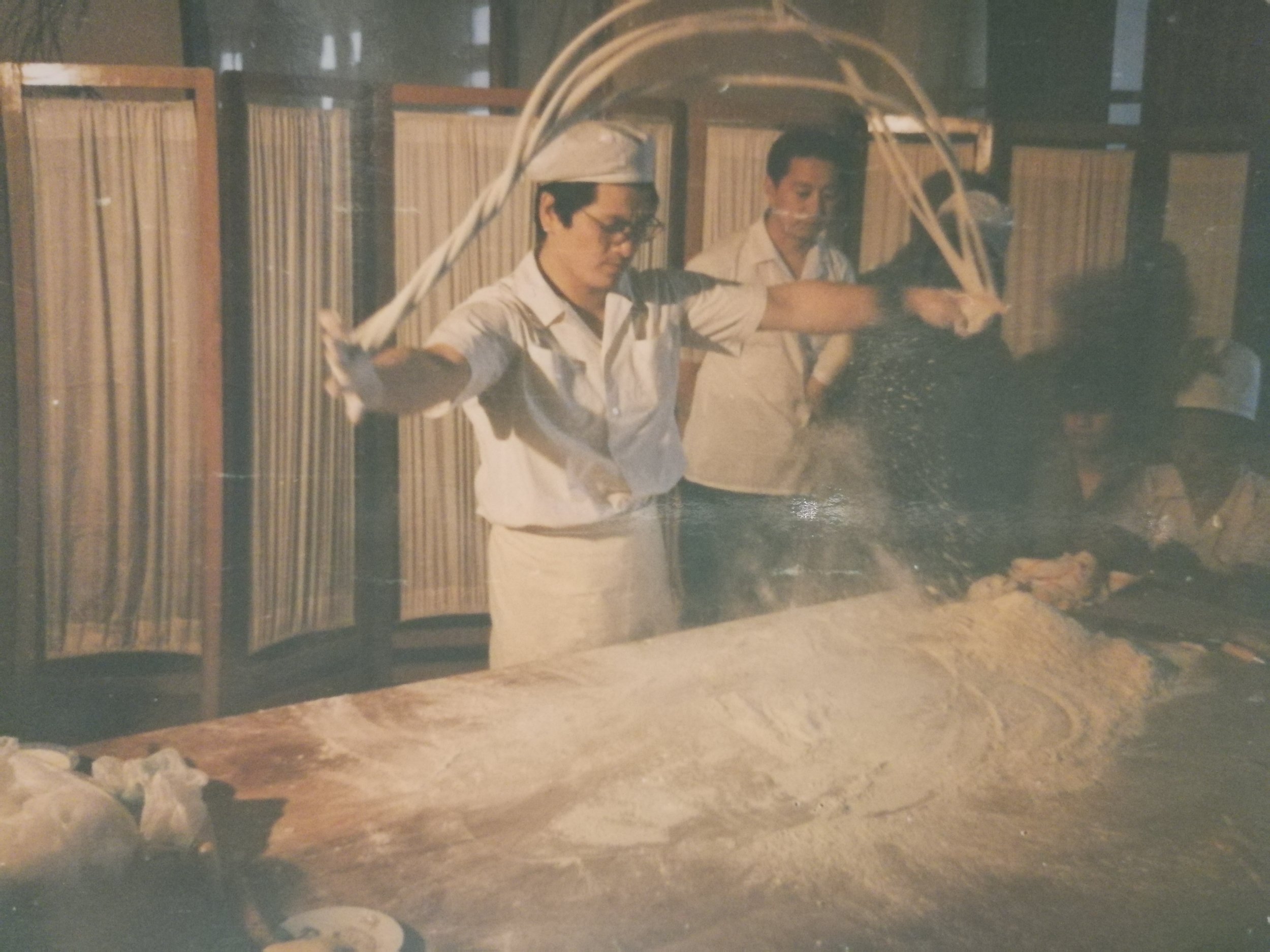A woman is swinging a whip like object over a table covered in flour, with steam or flour dust rising from the table, while a man watches nearby in a room with wooden room dividers.