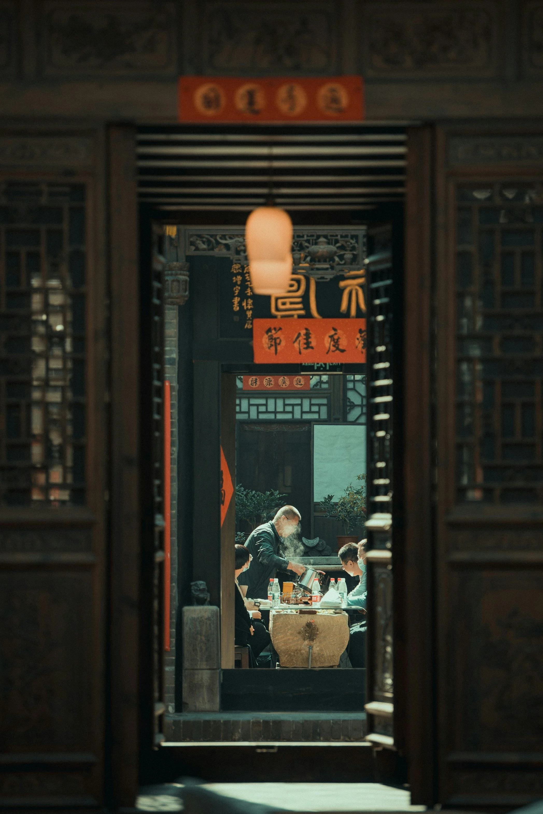 View through an ornate wooden doorway into a traditional Chinese restaurant where a waiter serves food to customers at a table, with decorative Chinese lanterns and calligraphy banners hanging.