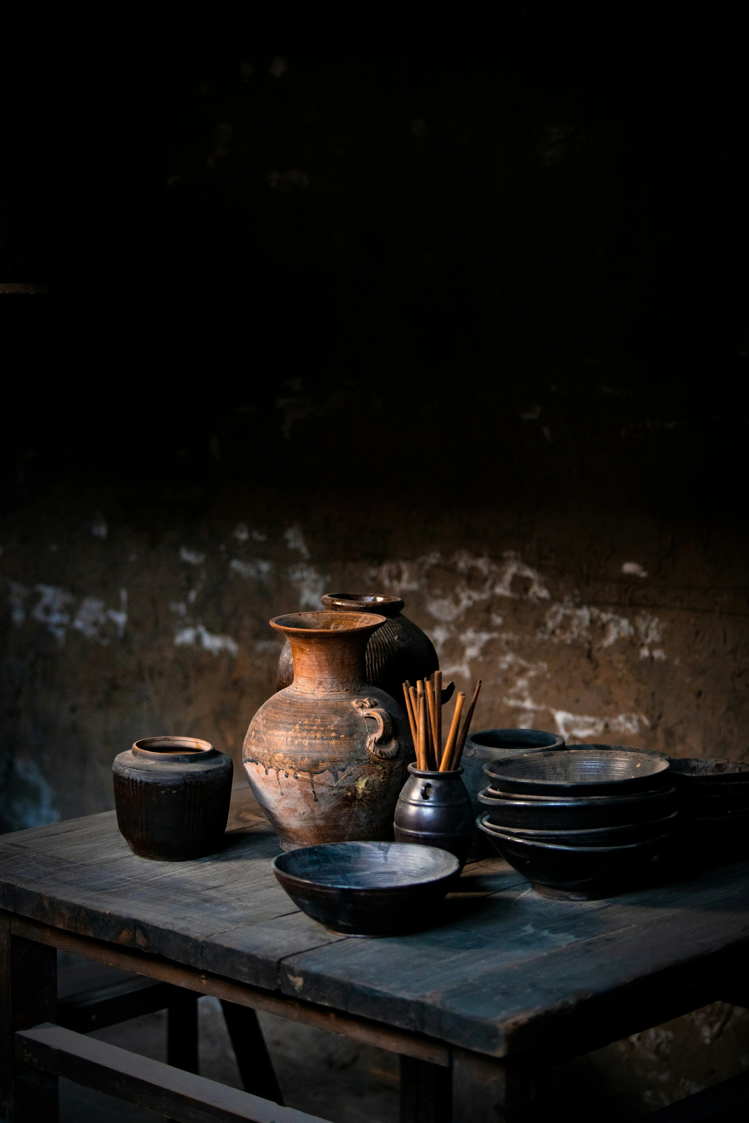 A rustic wooden table with various black and brown earthenware pots, bowls, and a small vase with wooden sticks inside, set against a dark, textured wall.