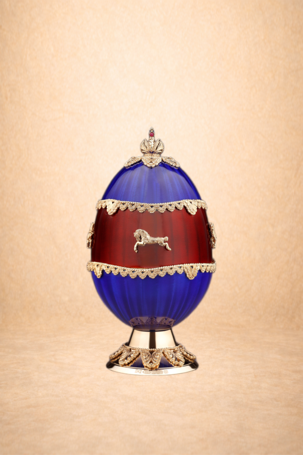 An ornate decorative egg with royal blue and red enamel, gold trim, and a silver lion emblem, set against a textured gold background.