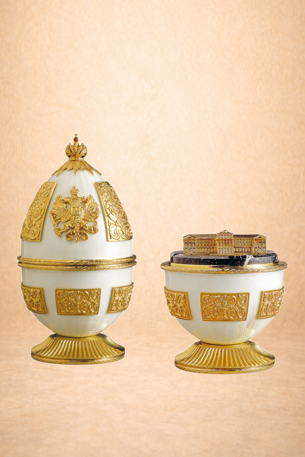 Decorative egg-shaped objects with gold embellishments, featuring a crest of a double-headed eagle on one and a building miniature on the other, set against a beige background.