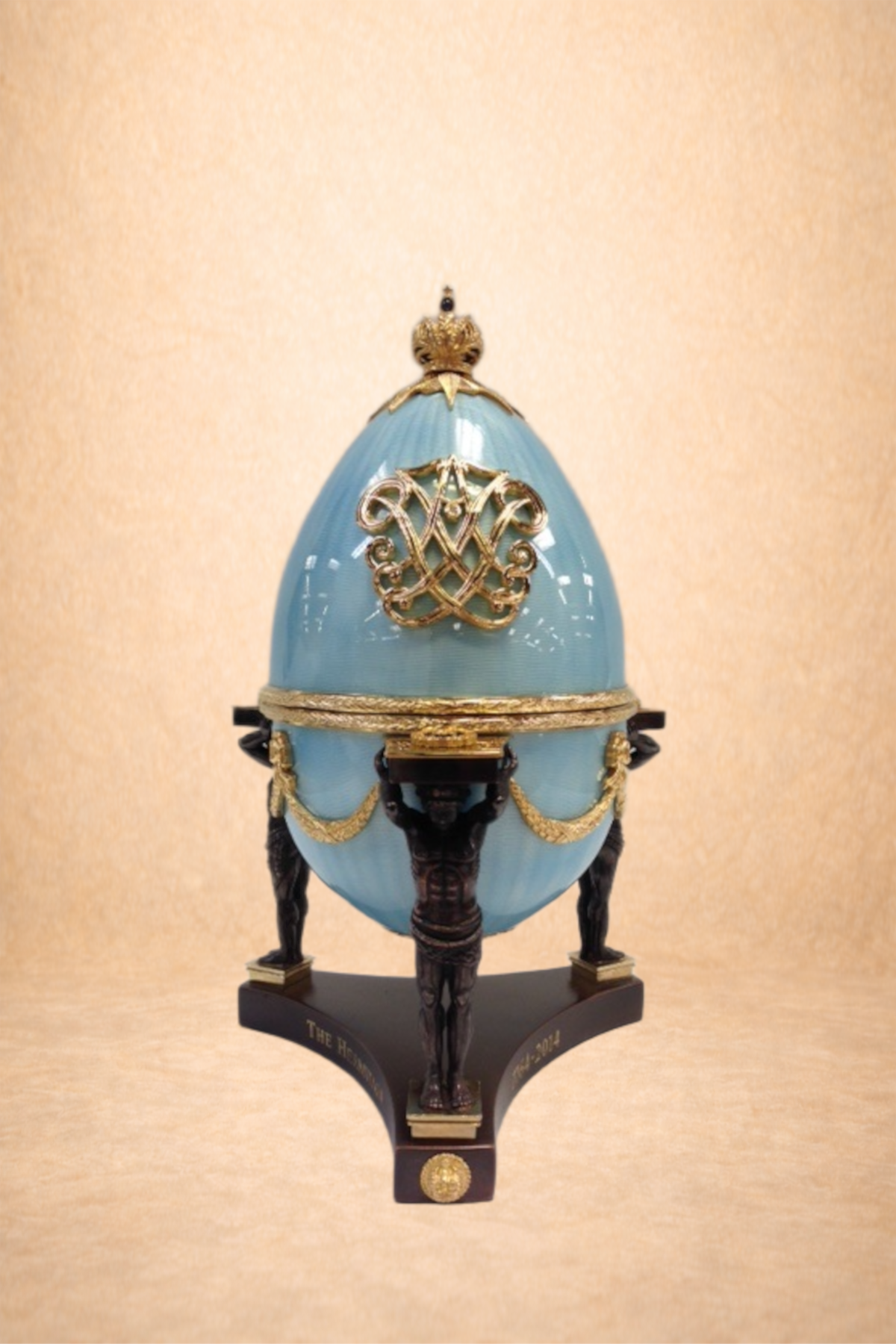 A decorative egg sculpture with black human figure legs, gold embellishments, a blue shell, and a gold crown on top, displayed on a black stand.