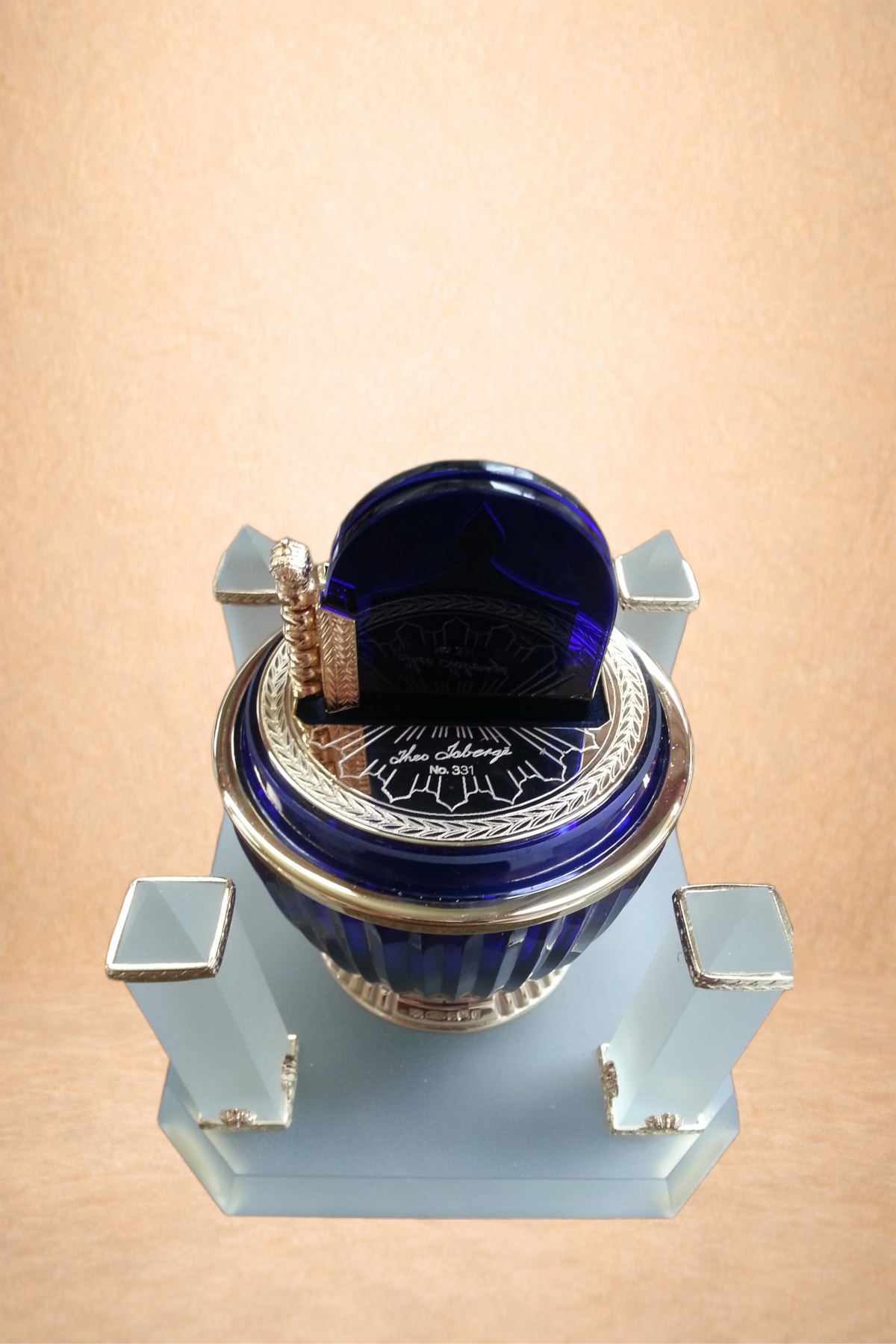 Top view of a decorative perfume bottle with a blue glass lid, silver accents, and a blue and silver base, placed on a light blue display stand.