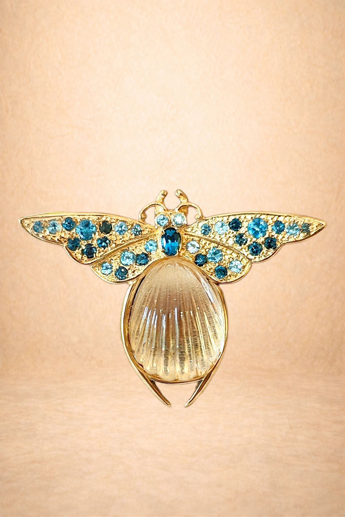 A gold brooch shaped like a bee with blue and clear gemstones on its wings and a pearl tail, set against a pink background.