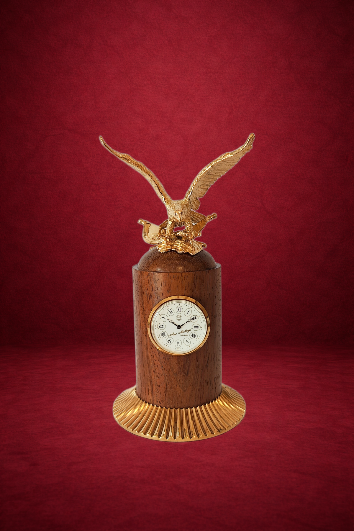 A decorative clock with a wooden base, gold accents, and a golden eagle sculpture on top, set against a red background.