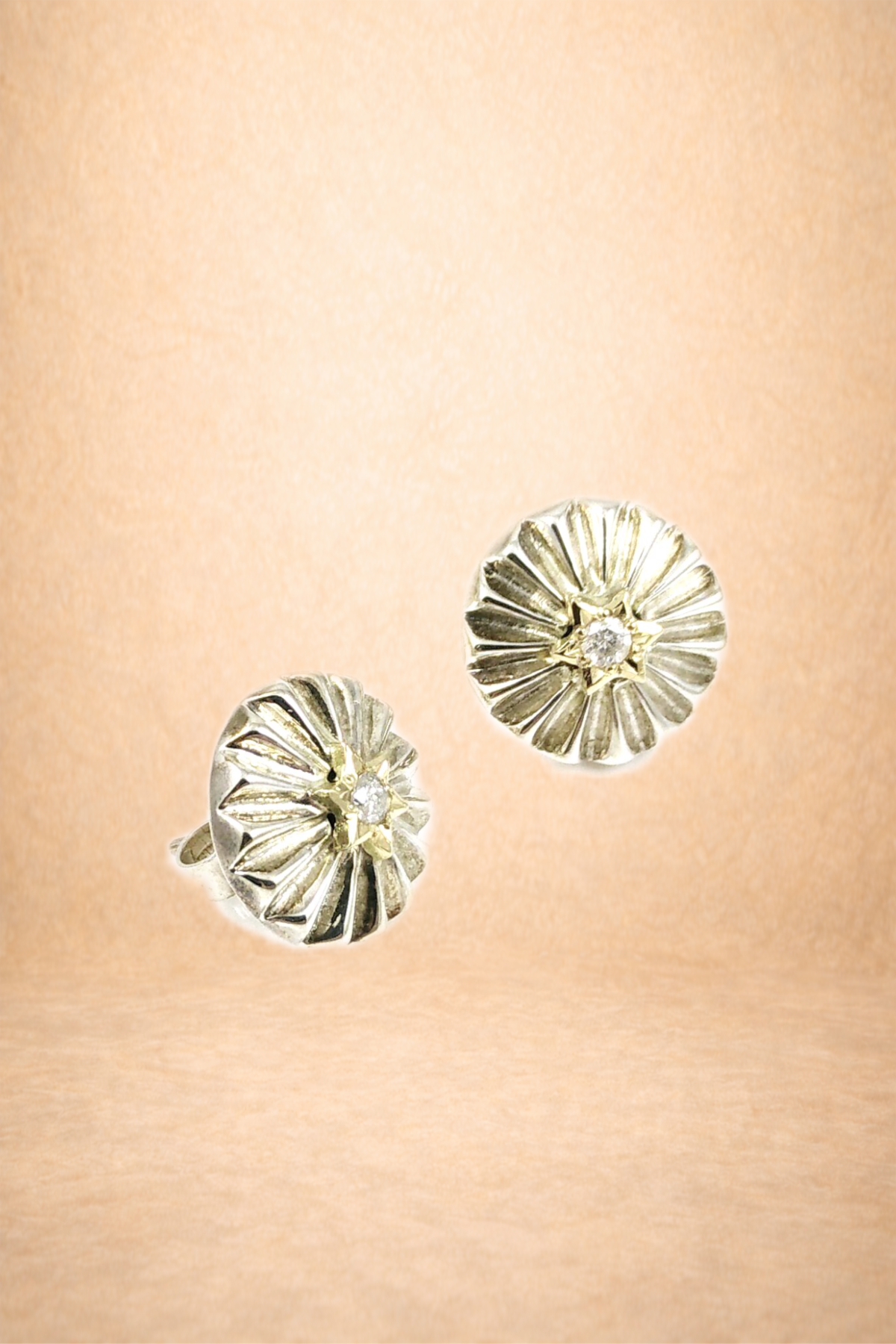 A close-up of a jewelry display showcasing a pair of gold and silver flower-shaped earrings with small diamonds in the center, against a warm, peach-colored background.
