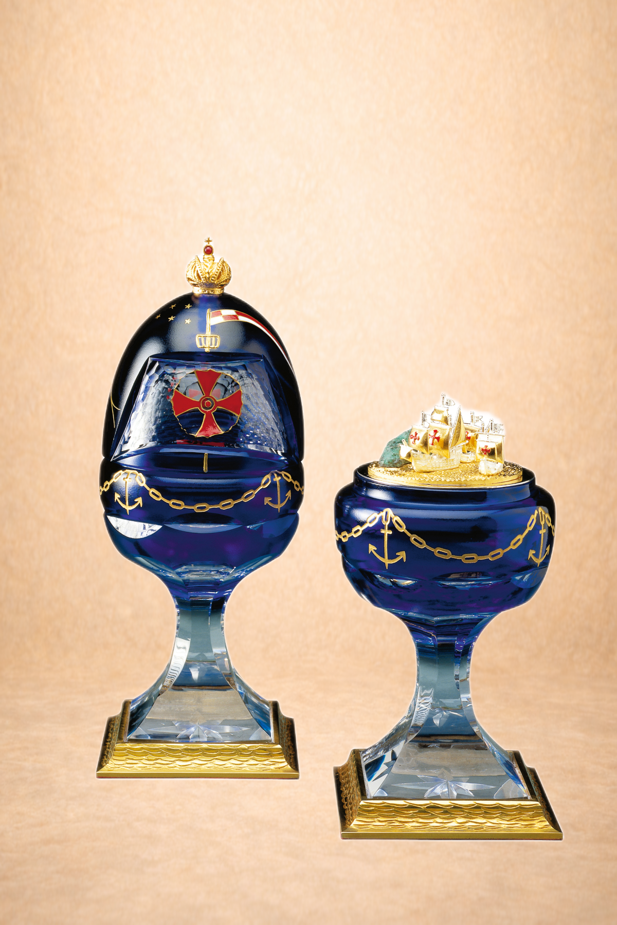 Two decorative glass items on glass pedestals, featuring navy blue color with gold anchor and chain motifs, and topped with royal and ship-themed ornaments.