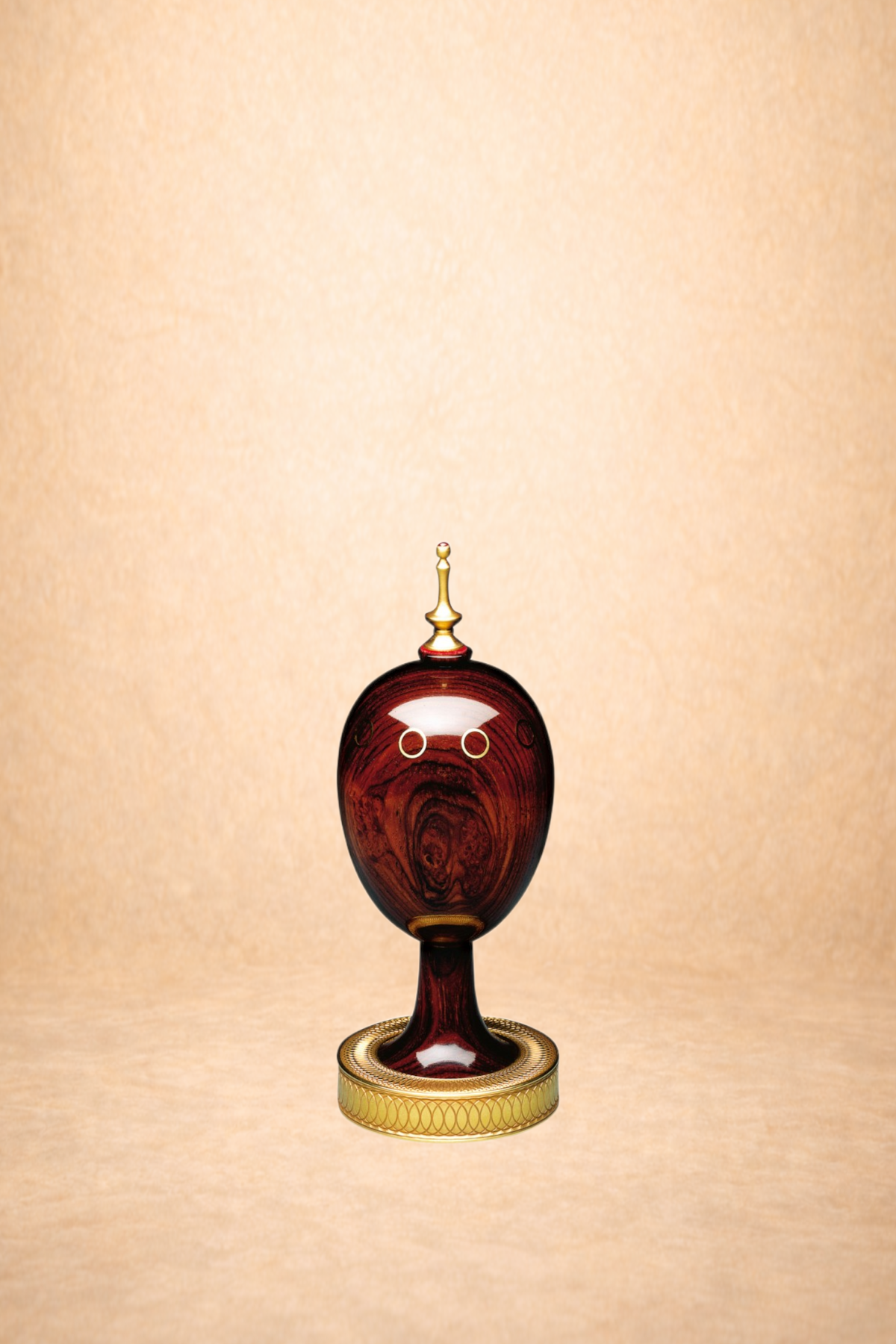 An ornate decorative object resembling a face with rings for eyes, a gold pointed ornament on top, and a circular base, placed against a beige background.