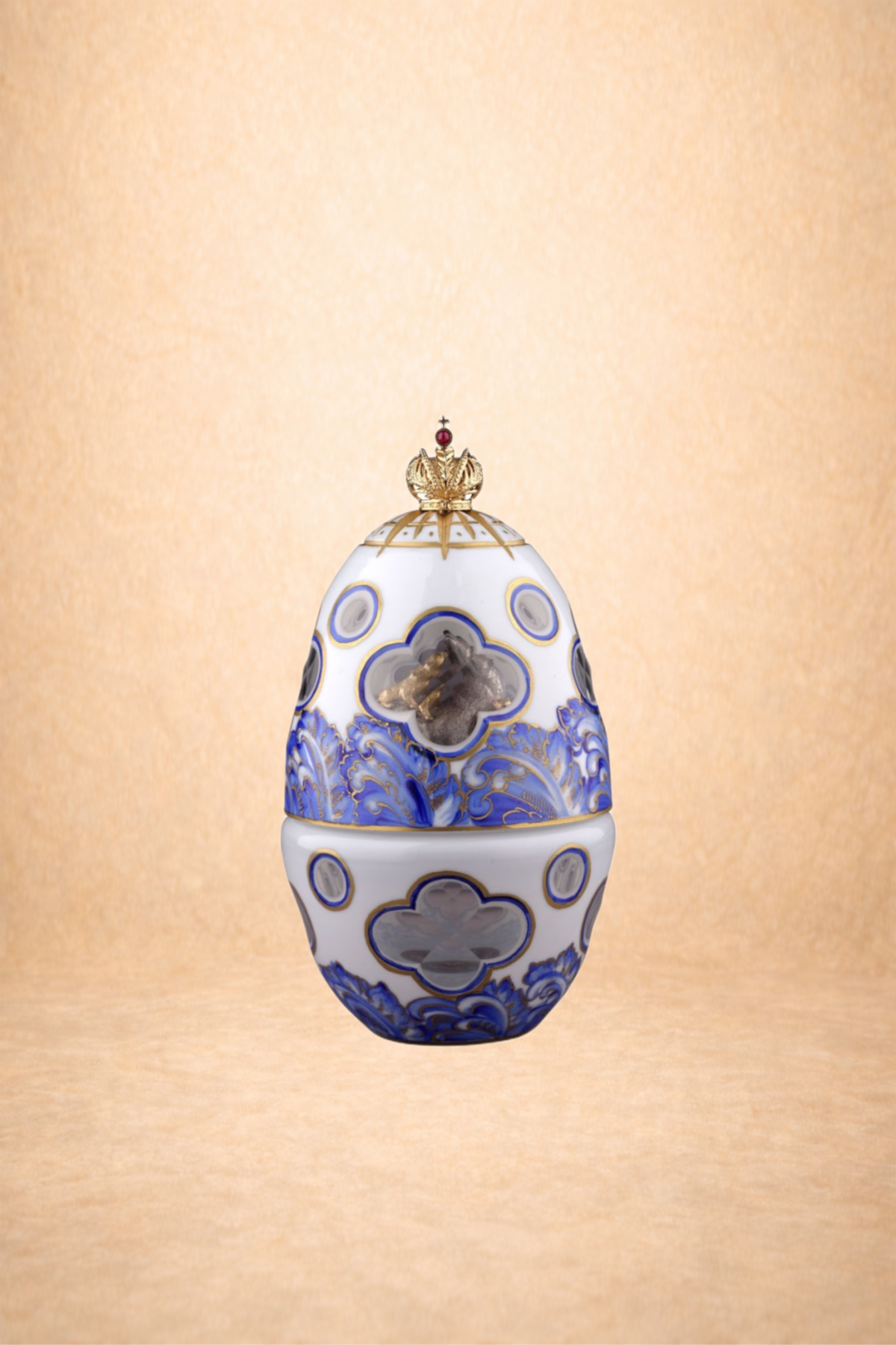A decorative porcelain egg with blue and gold designs, topped with a gold crown, and featuring some internal figures or objects visible through openings.
