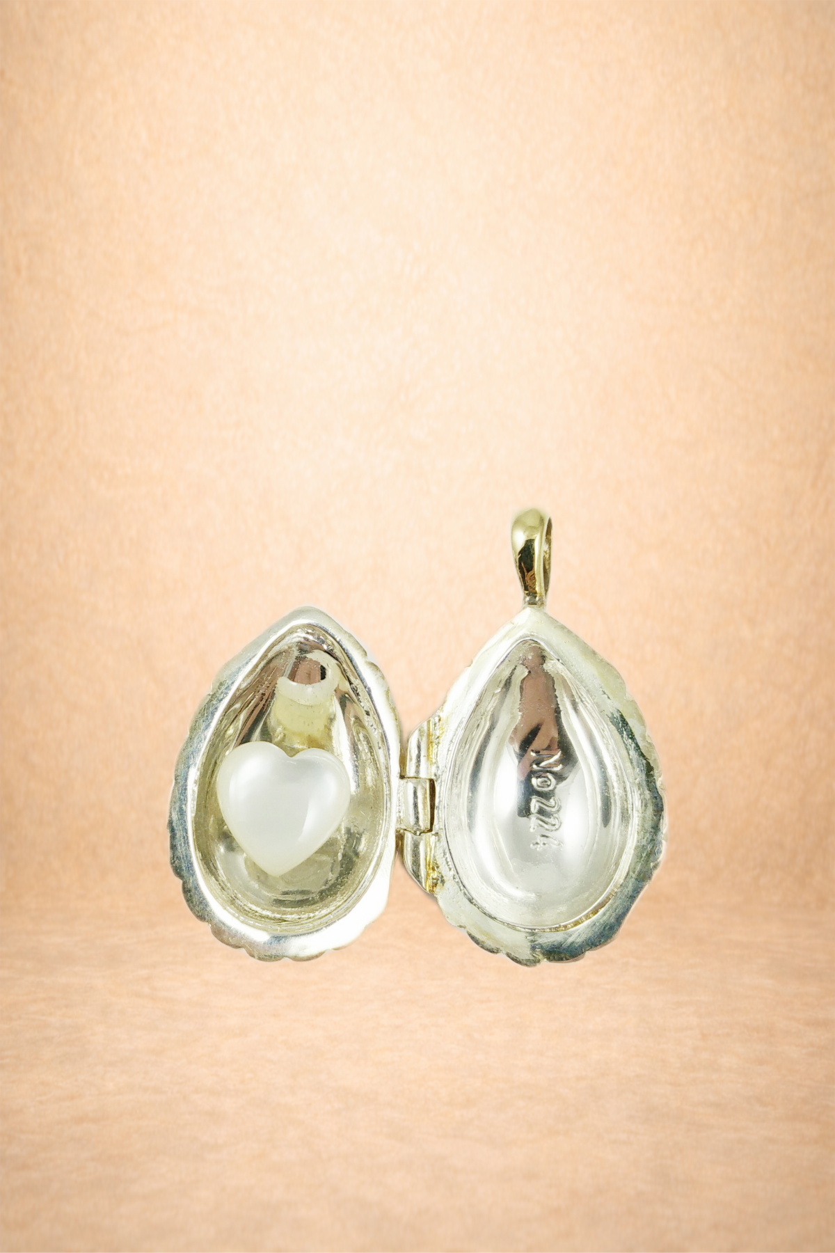 A silver jewelry box shaped like a clam shell, open, with a small pearl heart inside on the left side and a hollow interior on the right side, against a neutral background.