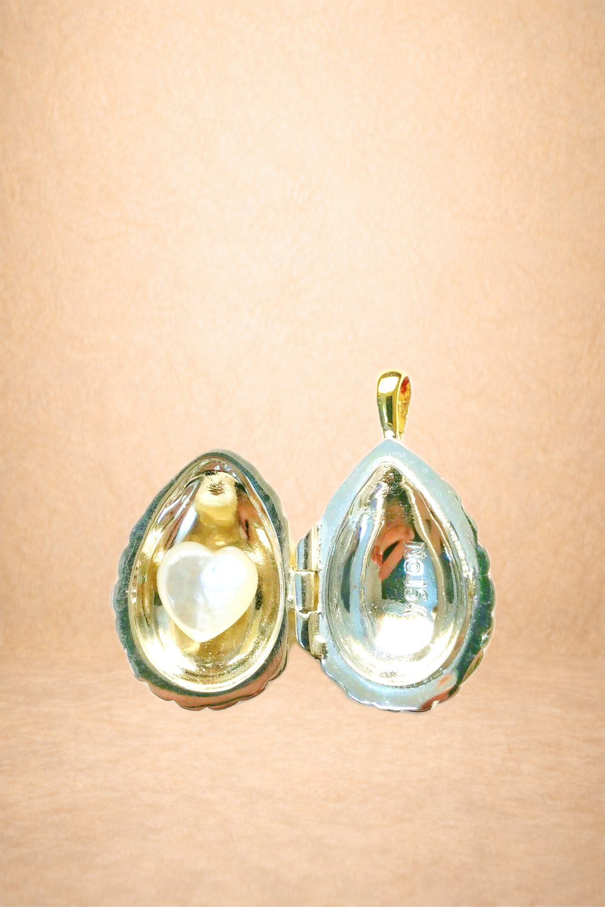 A gold jewelry locket shaped like an oyster shell, with a pearl in the shape of a heart inside, on a light pink background.