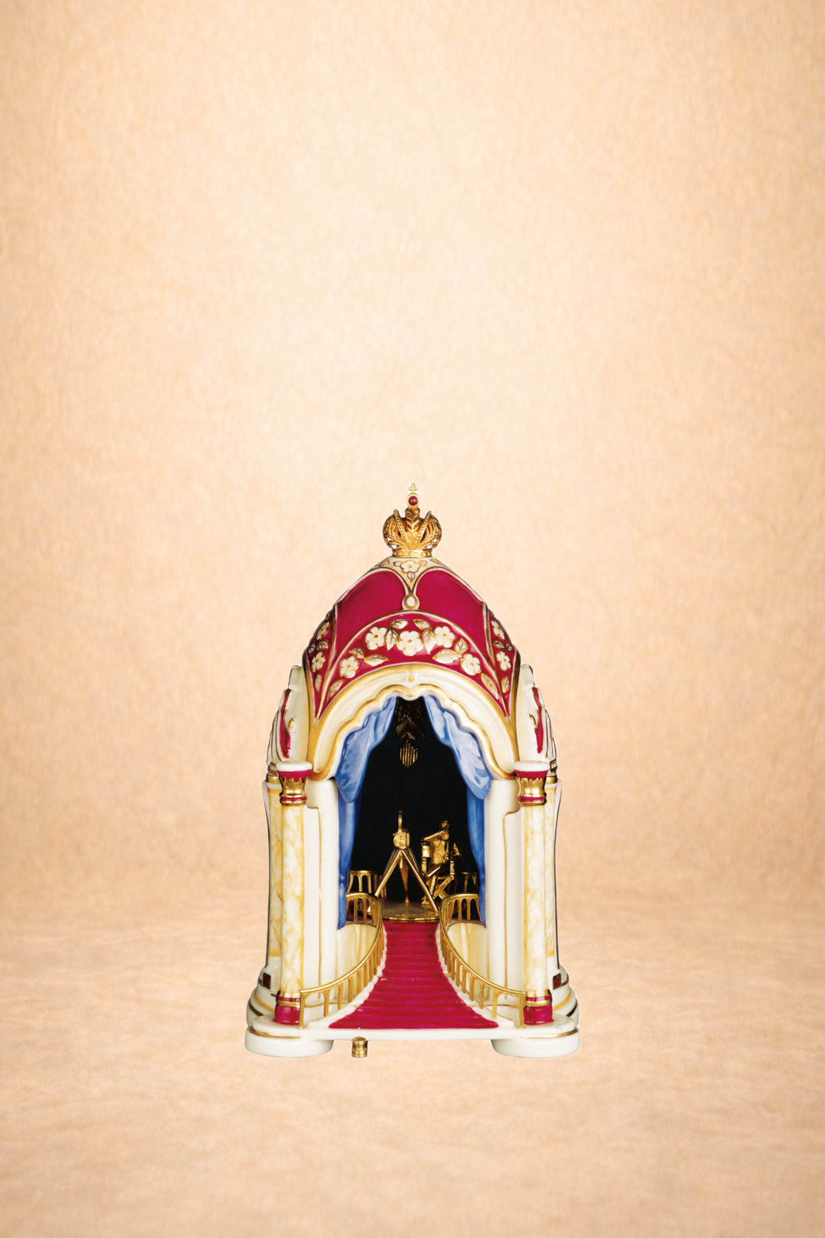 A miniature ornate church or castle with a red carpeted staircase leading to a black interior entrance, topped with gold and red accents, set against a beige background.