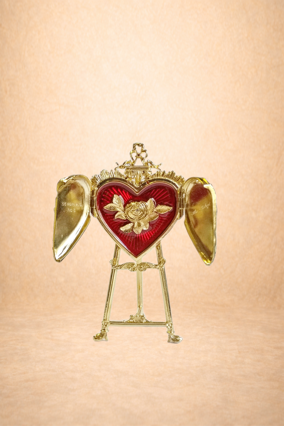 A gold picture frame with wings, shaped like a heart with a red background and gold rose design, standing on a stand against a beige background.