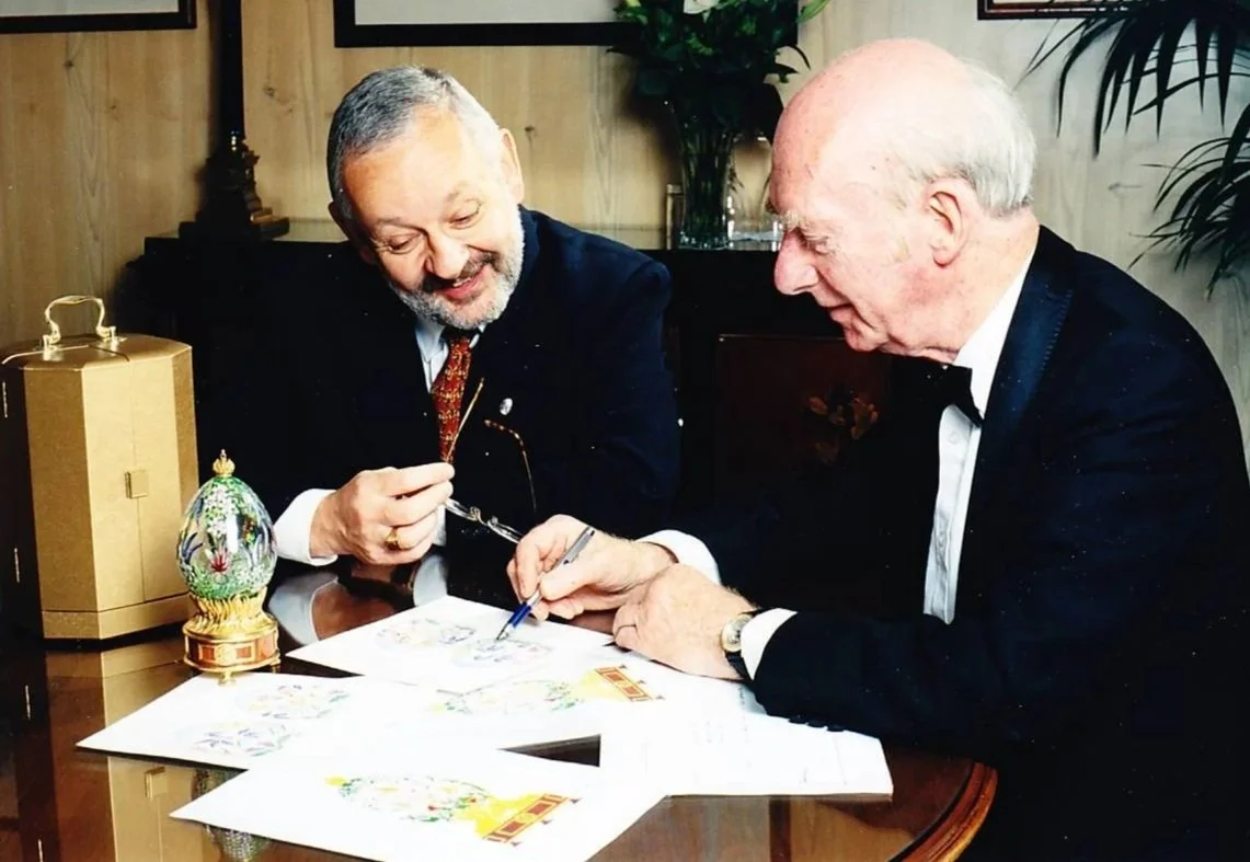 Two elderly men in tuxedos sitting at a table, examining and signing colorful artwork or cards.