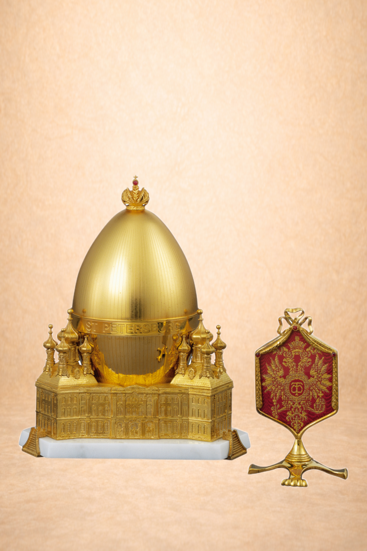 A golden replica of the St. Petersburg Winter Palace with a golden dome and small spires, and a red and gold emblem with the double-headed eagle on a black stand.