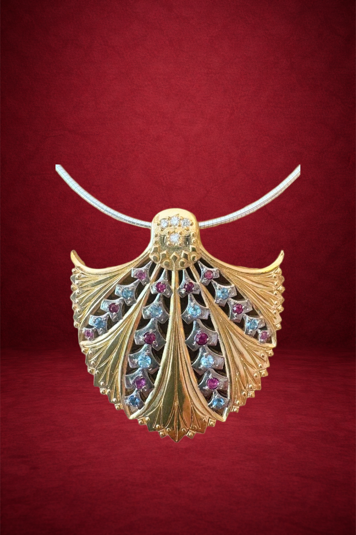 Gold leaf-shaped pendant with pink and clear gemstones on a silver chain, displayed against a red background.