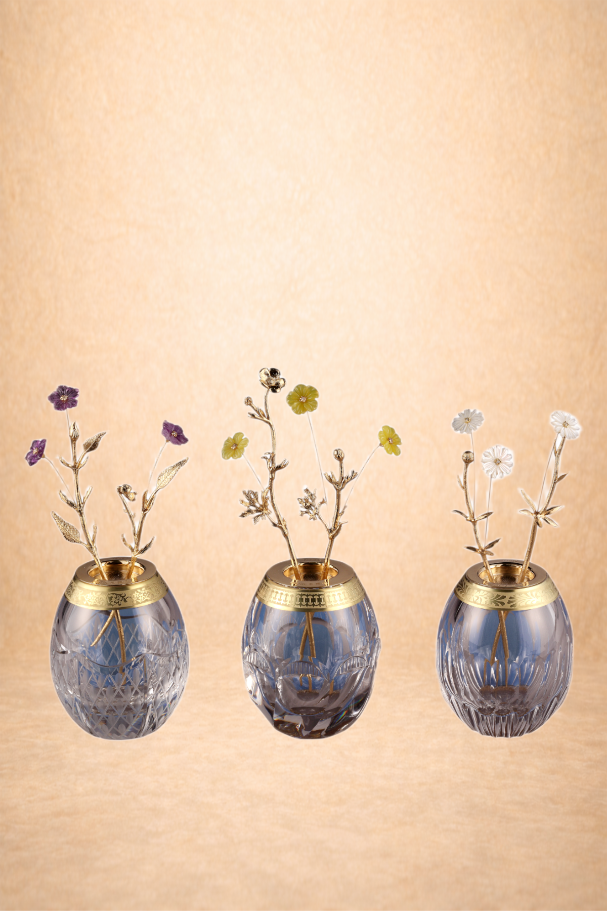 Three decorative glass vases with gold accents holding small artificial flowers, set against a beige background.