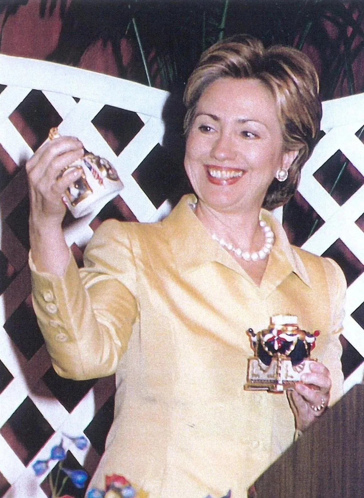 A woman with short brown hair wearing a gold blouse, pearl necklace, and pearl earrings, smiling and holding a small porcelain centerpiece decorated with miniature American flags and bunting, sitting on a white lattice or fence with a tropical or garden background.