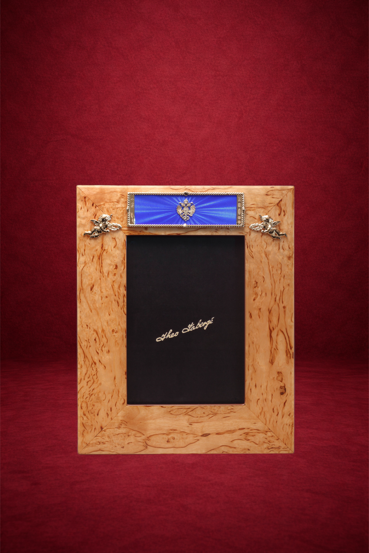 A wooden picture frame on a red background with a small blue ribbon and two silver angel ornaments at the top, and a black photo mat with white cursive text reading 'Ahea Hoberge' in the center.