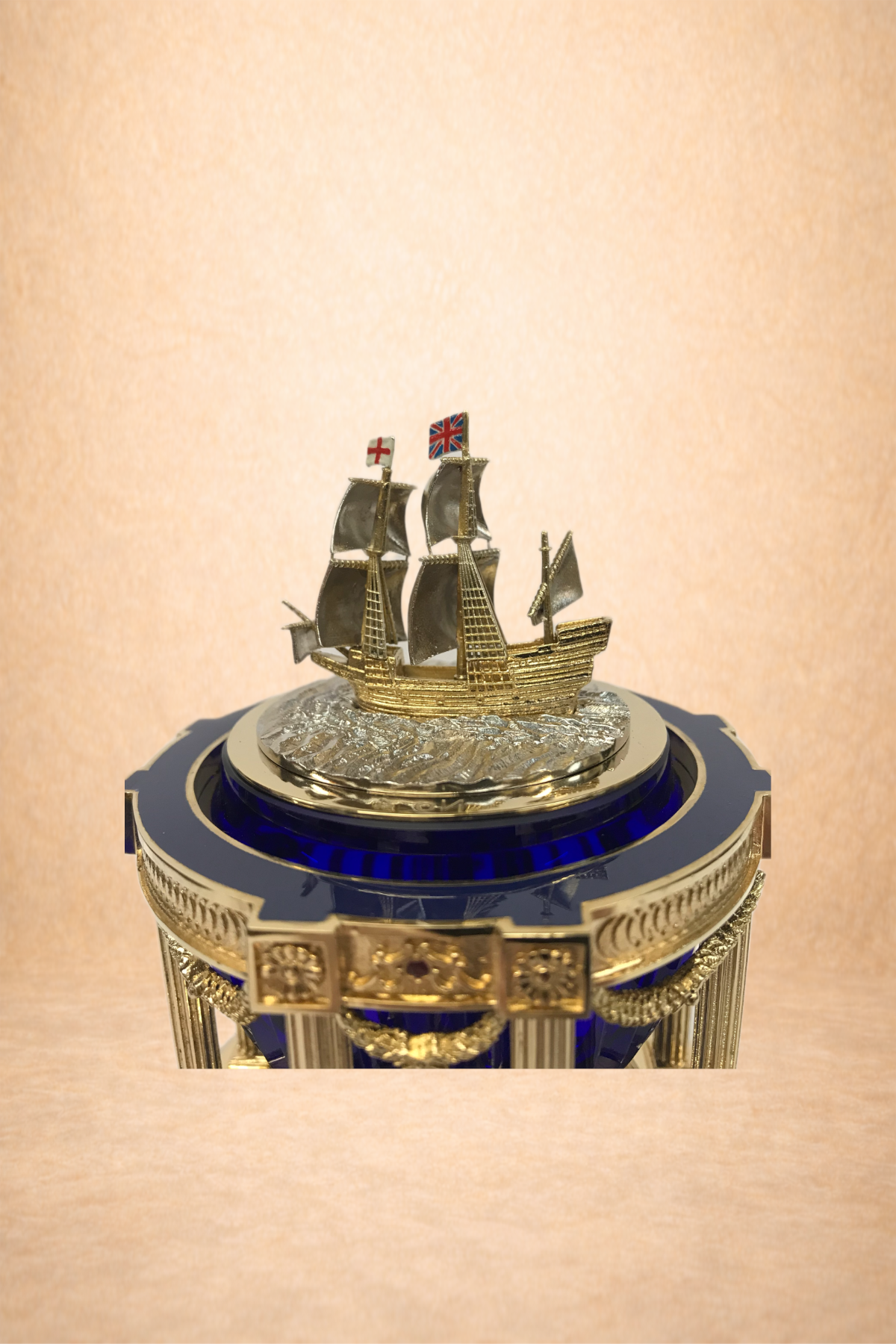A detailed miniature model of an old sailing ship with three masts, each flag showing the Union Jack and St. George's Cross. The ship is displayed on a decorative blue and gold stand with ornate embellishments.