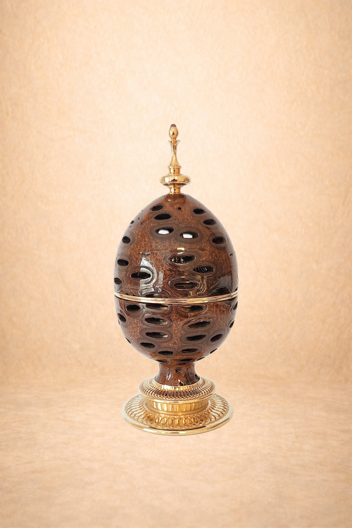 Decorative oval container with a perforated brown exterior, topped with a gold finial, and designed to resemble an Easter egg, standing on a gold decorative base.