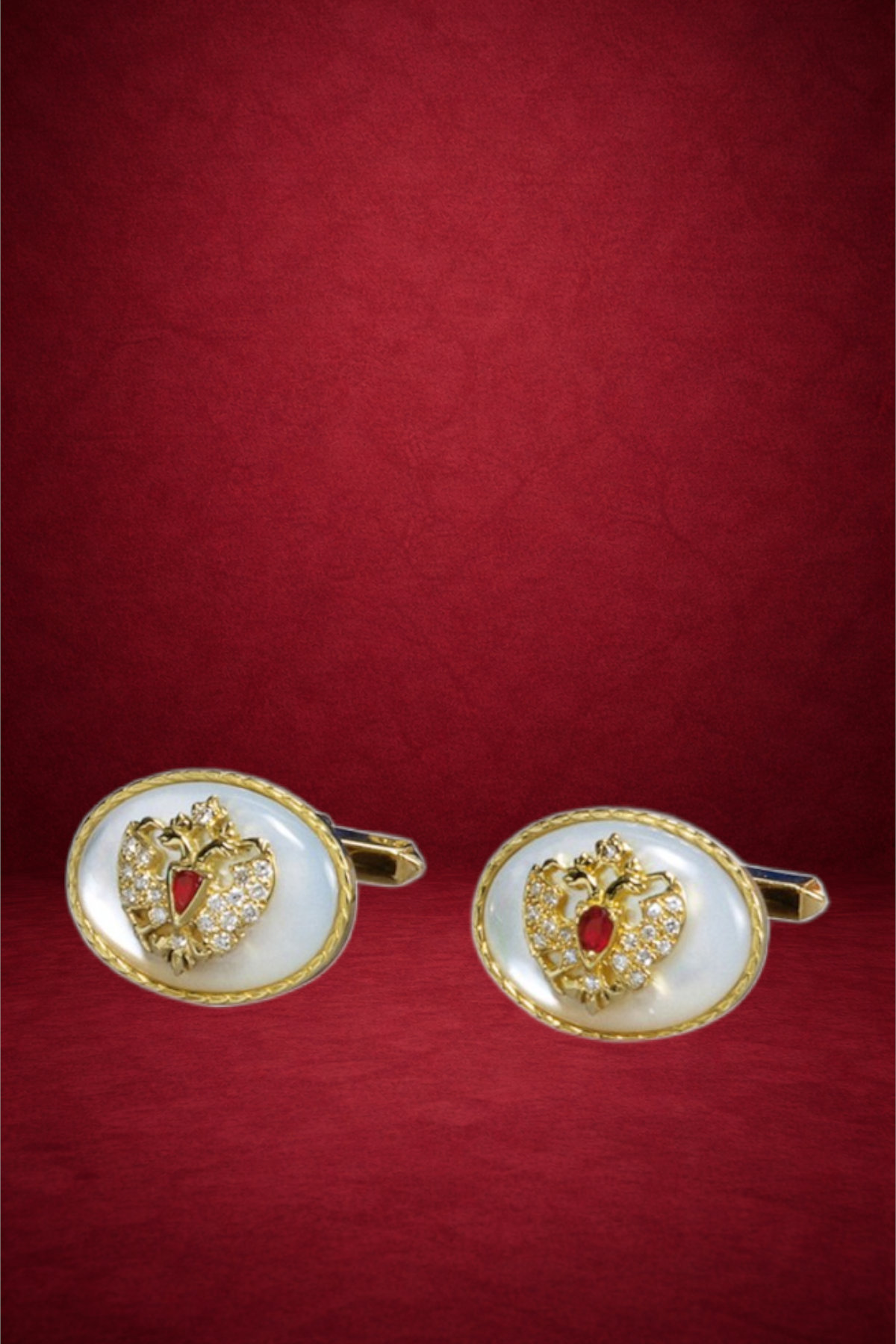 Pair of gold and pearl cufflinks with heart-shaped designs and red gemstones, set against a red background.
