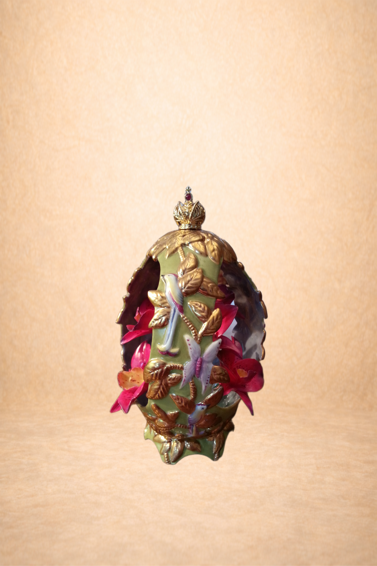 Decorative oval-shaped object with intricate floral and bird design, adorned with gold accents, red flowers, and a small crown-like top, placed on a plain background.