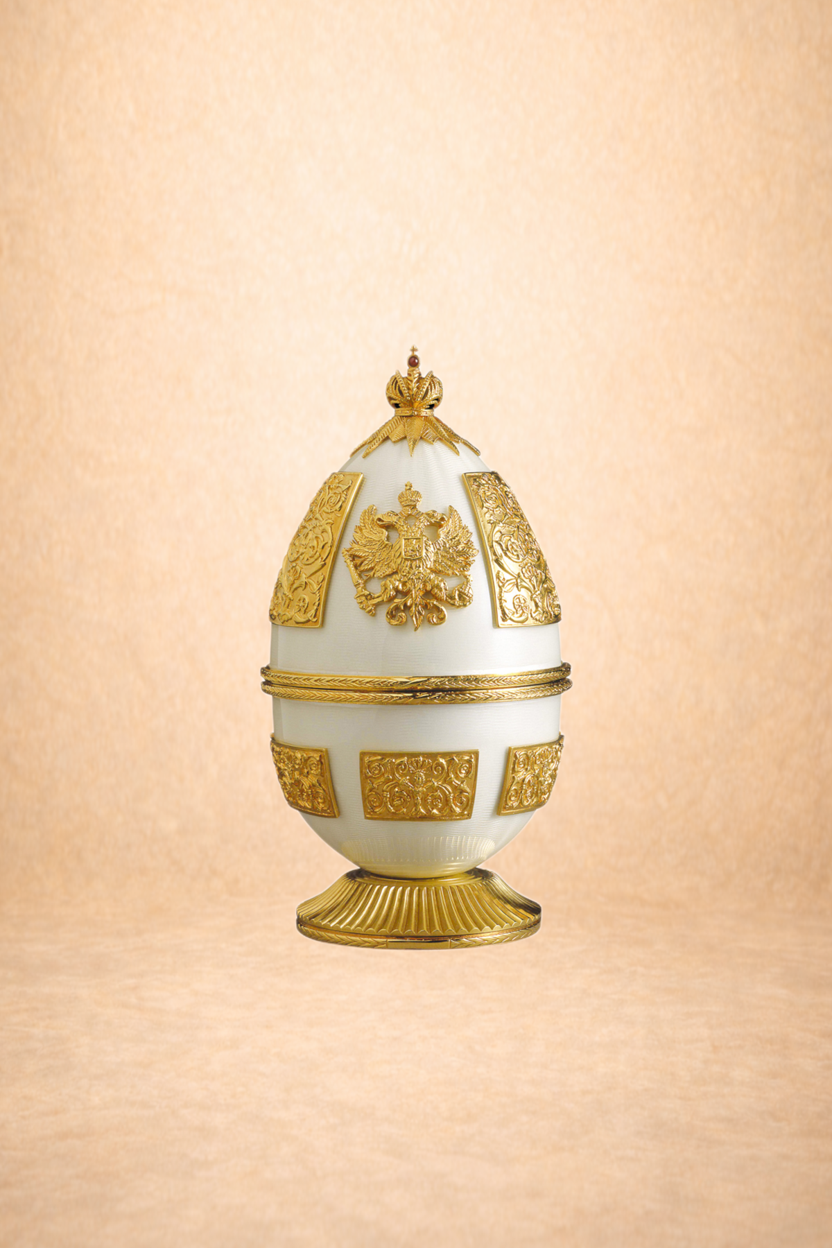 A decorative egg with a white body and gold accents, featuring intricate gold reliefs and the Russian coat of arms, set against a beige background.